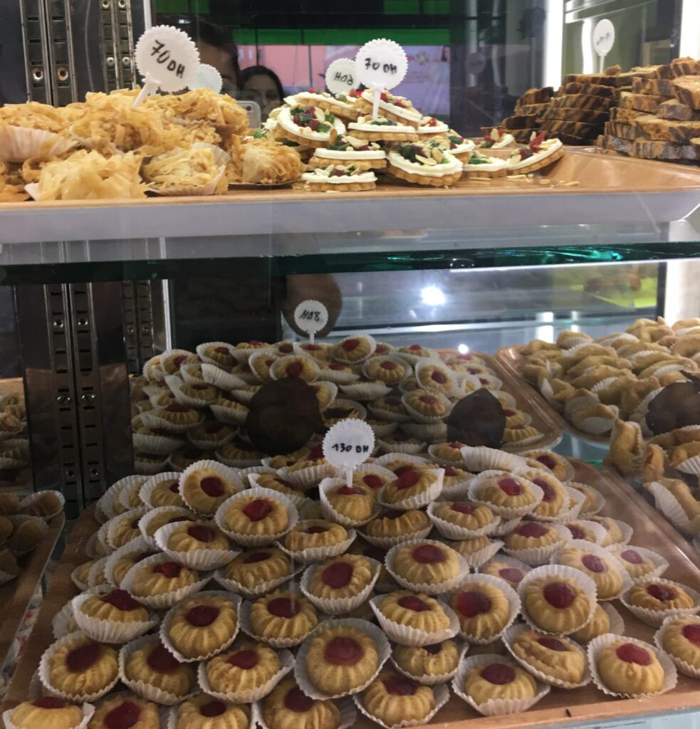A bakery display filled with rows of pastries behind glass, with two people reflected in the cabinet as they look at the choices.